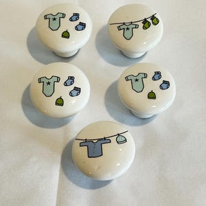 May include: Five round white ceramic cabinet knobs featuring hand-painted illustrations of baby clothing. The designs include light blue onesies, small socks, and green hats, with some items depicted hanging on a clothesline and others floating freely.
