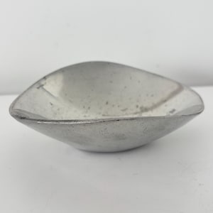 May include: A small, silver-coloured, leaf-shaped bowl. The bowl has a smooth, slightly textured surface and a rounded, asymmetrical shape. The bowl is empty and sits on a white surface.