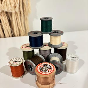 Könnte beinhalten: Eine Sammlung von Vintage-Garnrollen in verschiedenen Farben, darunter Grün, Marineblau und Braun. Die Rollen sind auf einer weißen Oberfläche angeordnet, wobei einige zu einer kleinen Pyramide gestapelt sind. Die Etiketten auf den Rollen sind sichtbar.