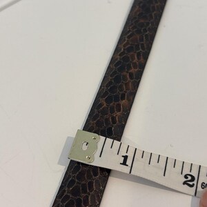 May include: A brown leather strap with a textured pattern is measured with a white measuring tape. The tape shows the strap is approximately 1 inch wide. The strap has a dark brown and black color scheme.
