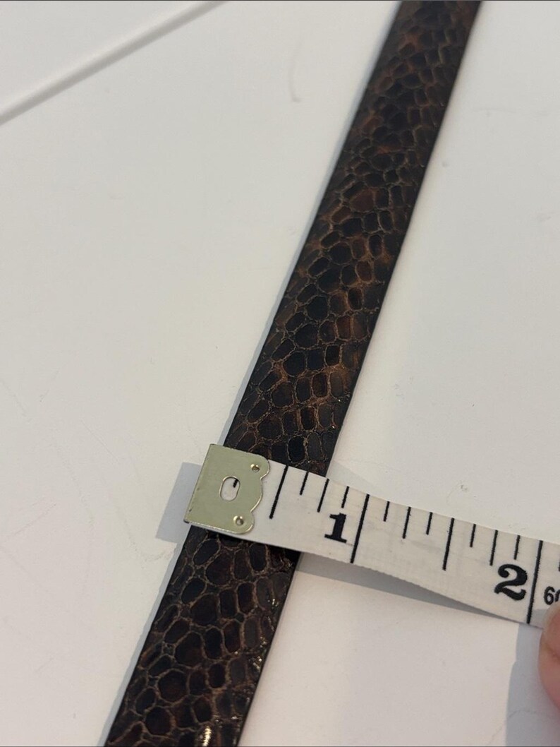 May include: A brown leather strap with a textured pattern is measured with a white measuring tape. The tape shows the strap is approximately 1 inch wide. The strap has a dark brown and black color scheme.