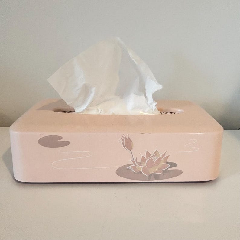 May include: A peach-coloured rectangular tissue box with a floral lotus design. White tissues are pulled from the top. The box is a decorative item for home use. The design includes a lotus flower and lily pads.