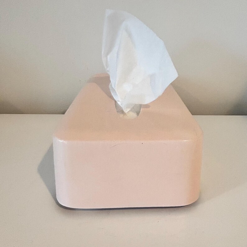 May include: A light pink tissue box with a rectangular shape and rounded corners. White tissues protrude from the top. The box is made of a smooth material and is placed on a white surface.