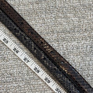 May include: Two snake-skin patterned belts in shades of brown and black, with a measuring tape showing lengths from 25 to 31 inches. The belts have small holes for the buckle and a textured surface.