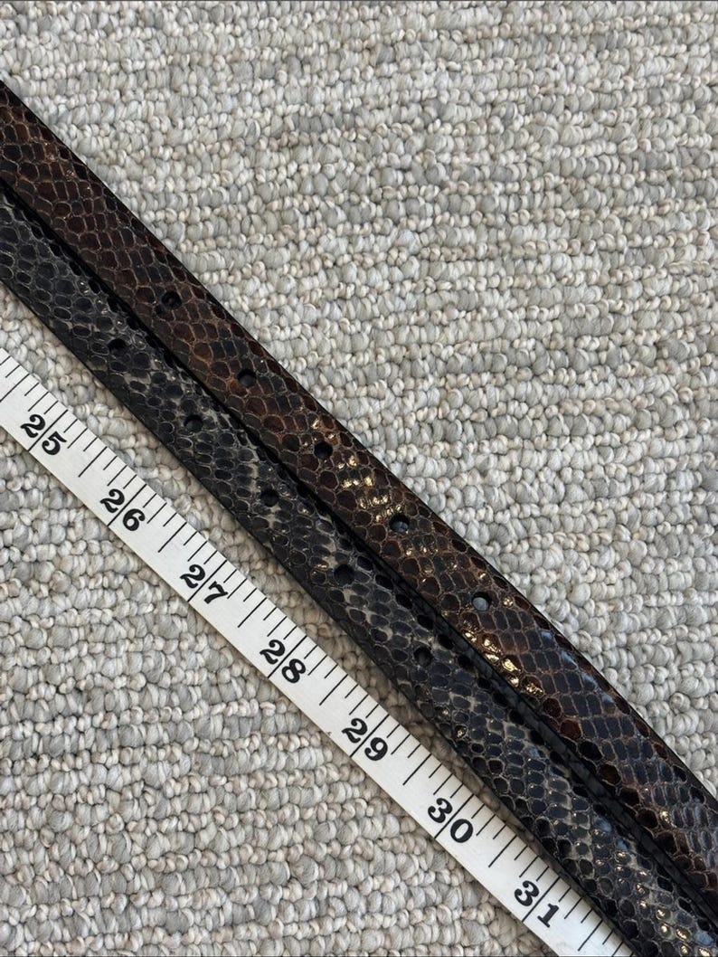 May include: Two snake-skin patterned belts in shades of brown and black, with a measuring tape showing lengths from 25 to 31 inches. The belts have small holes for the buckle and a textured surface.