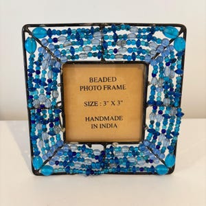 May include: Square beaded photo frame with a black metal frame. The frame is decorated with blue and turquoise beads. The text on the frame reads: "BEADED PHOTO FRAME SIZE: 3" X 3" HANDMADE IN INDIA."