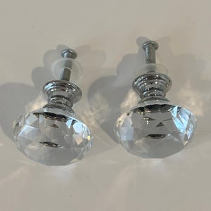 Könnte beinhalten: Zwei klare Kristall-Schrankknöpfe mit facettierten Oberflächen und verchromten Metallsockeln. Die Knöpfe haben ein klassisches, elegantes Design, perfekt, um Möbeln oder Schränken einen Hauch von Glanz zu verleihen. Jeder Knopf hat eine Schraube für eine einfache Installation.