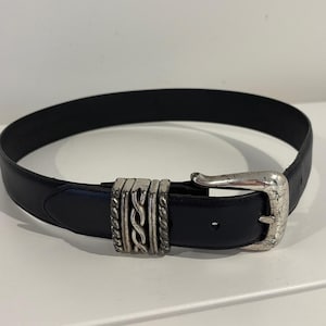 May include: A black leather belt featuring a silver-toned metal buckle and a decorative sliding keeper with a twisted rope design. The buckle has a weathered, textured finish, and the belt strap includes a single visible adjustment hole.