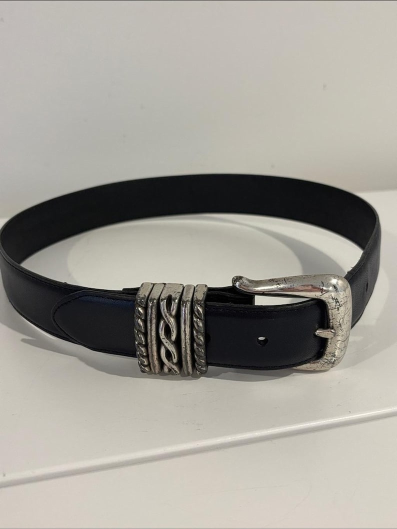 May include: A black leather belt featuring a silver-toned metal buckle and a decorative sliding keeper with a twisted rope design. The buckle has a weathered, textured finish, and the belt strap includes a single visible adjustment hole.