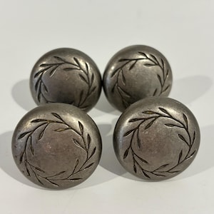 May include: Four antique silver-toned cabinet knobs. Each knob is round with a raised leaf design around the edge. The knobs are made of metal and have a slightly tarnished appearance, suggesting age.