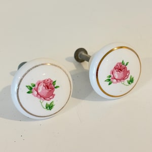 May include: Two white ceramic cabinet knobs with a pink rose design and gold trim. The knobs have a metal stem for mounting. The floral design includes green leaves. These decorative knobs are ideal for furniture or cabinet upgrades.