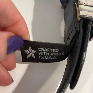 May include: A black belt featuring a rectangular silver-toned metal buckle with a distressed finish. A black fabric tag is attached to the belt, displaying a white star icon and the text CRAFTED WITH PRIDE IN U.S.A. in white capital letters.