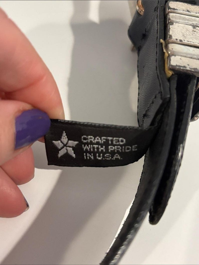 May include: A black belt featuring a rectangular silver-toned metal buckle with a distressed finish. A black fabric tag is attached to the belt, displaying a white star icon and the text CRAFTED WITH PRIDE IN U.S.A. in white capital letters.