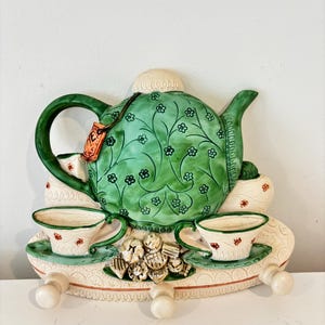 Op de afbeelding: Een keramische wand-theeset met een groene theepot met bloemendetails en twee bijpassende kopjes. De set bevat een decoratief dienblad met drie haken om op te hangen. De theepot heeft een label met "Tea".