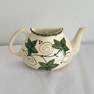 May include: A cream-coloured ceramic wall pocket shaped like a teapot, featuring a hand-painted green ivy leaf pattern with swirling vines and small dots. The piece has a flat back with a small hole for hanging, a curved handle, and a spout.