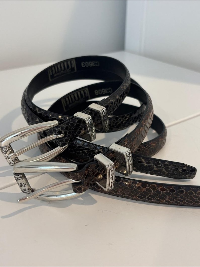 May include: Three brown and black patterned belts with silver-colored buckles. The belts are coiled and stacked, showcasing a textured, snake-like design. The buckles have an ornate design. The text "CLASSICS" and "C3603" is visible on the black belt.