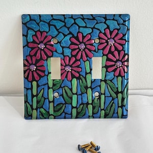 May include: A double light switch wall plate featuring a mosaic-style pattern of pink flowers with purple centres against a blue and black background, accompanied by four small brass-coloured metal screws for installation purposes.