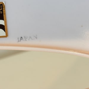 May include: A close-up of a pale pink dish with the word "JAPAN" printed in grey. A gold and brown sticker is visible in the upper left corner. The dish has a curved edge and a light beige background.