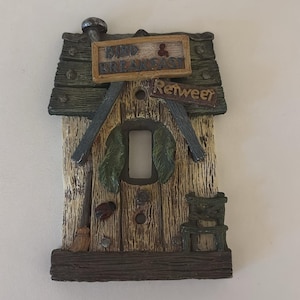 May include: Decorative light switch cover in the shape of a birdhouse. The cover is made of wood-like material with a weathered appearance. It features the words "Bird & Breakfast" and "Retweer". The design includes a small broom, chair, and a ladybug.
