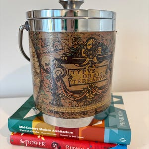 May include: A vintage ice bucket with a map design and the text "TYPVS ORBIS TERRARVM". The bucket has a silver lid and handle. It rests on a stack of books, including "Mid-Century Modern Architecture" and "the POWER".