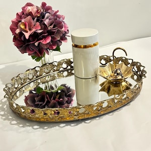 Puede incluir: Una bandeja ovalada con espejo y borde de metal dorado adornado con recortes florales. Sobre la bandeja hay un frasco cilíndrico blanco con una banda dorada, un pequeño plato dorado en forma de paraguas con joyas y un florero de cristal.