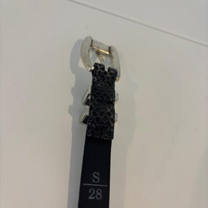 May include: Black belt with a silver buckle and a textured snakeskin pattern. The belt is labeled with the size "S" and the number "28".