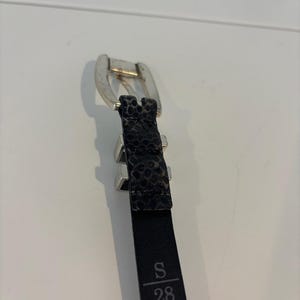 May include: Black belt with a silver buckle and a textured, patterned design. The belt is labeled with "S" and "28", indicating a size of 28 inches.