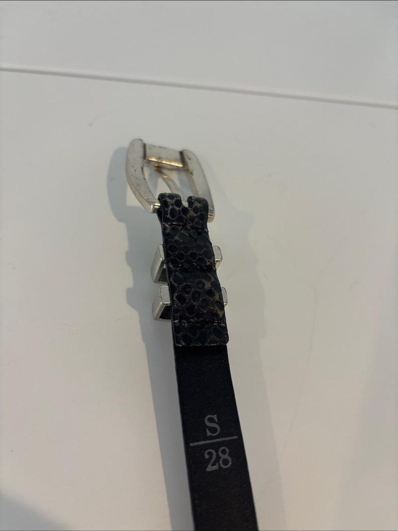 May include: Black belt with a silver buckle and a textured, patterned design. The belt is labeled with "S" and "28", indicating a size of 28 inches.