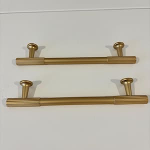 Op de afbeelding: Twee goudkleurige kastgrepen. Elke greep heeft een cilindrische staaf met geribbelde details aan de uiteinden en een afgeronde knop. De grepen zijn ontworpen voor een gemakkelijke grip.