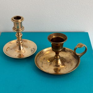 Puede incluir: Dos candelabros de latón con acabado metálico dorado. Uno presenta un tallo alto y delgado con base circular, mientras que el otro es un modelo más corto y ancho con una pequeña asa lateral unida a la base circular.