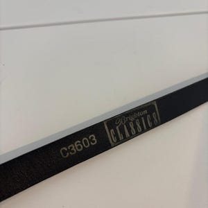 May include: A close-up of a black leather belt with the text "C3603" and "Brighton CLASSICS" printed on it. The belt is lying on a white surface.