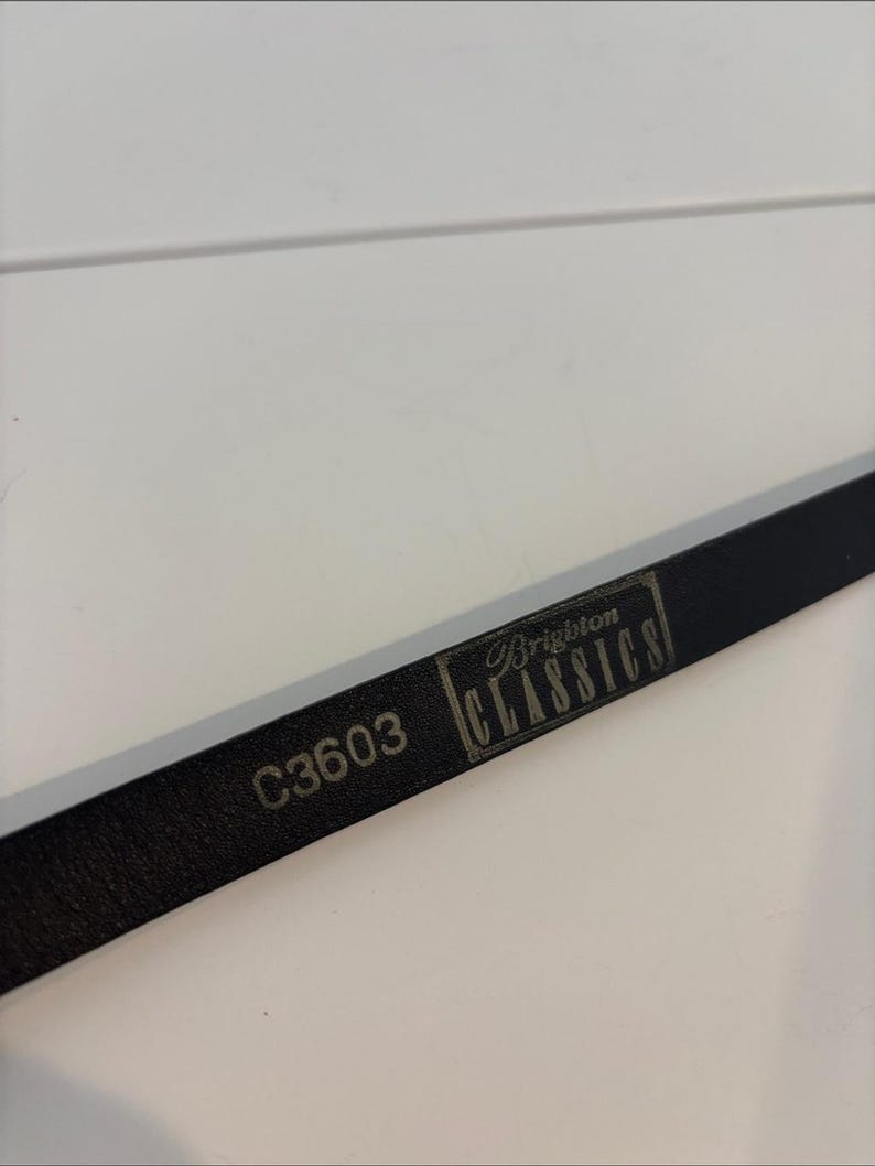 May include: A close-up of a black leather belt with the text "C3603" and "Brighton CLASSICS" printed on it. The belt is lying on a white surface.