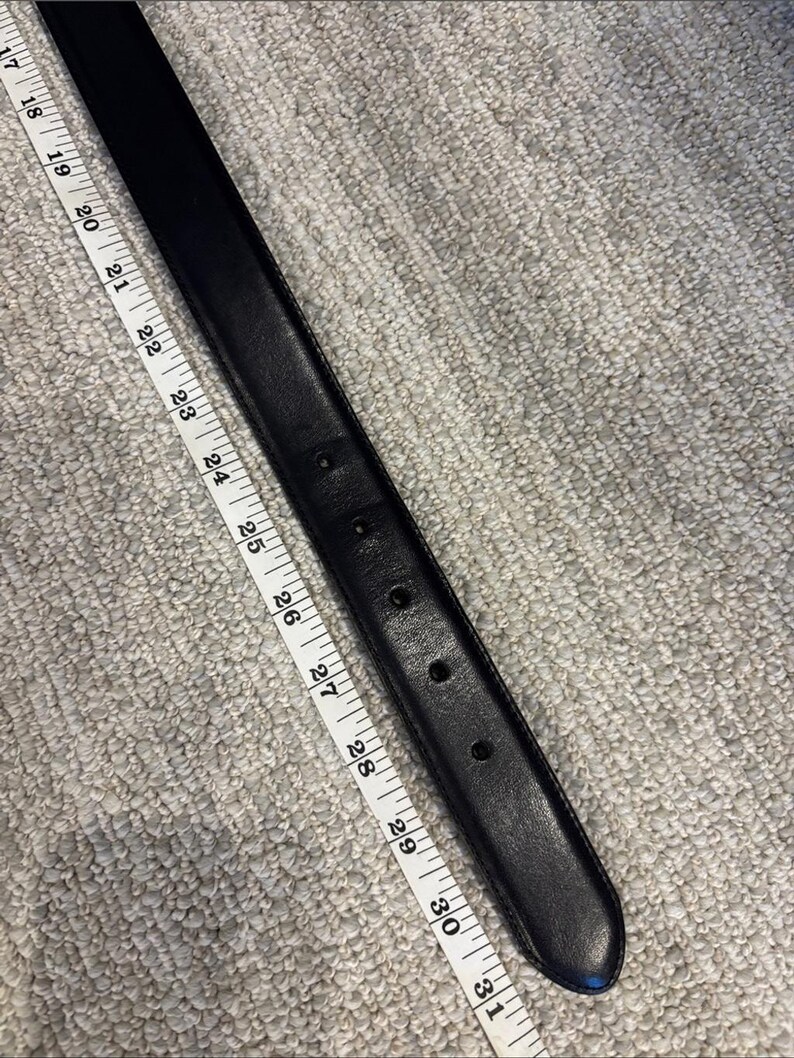 May include: A black belt with a series of adjustment holes, positioned alongside a white measuring tape that indicates a length measurement extending from 17 inches to 31 inches along the strap of the belt.