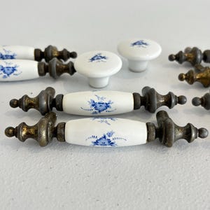 May include: A collection of white ceramic drawer pulls and knobs featuring blue floral patterns. The hardware includes elongated handles with metallic, aged-finish ends and round knobs, all decorated with delicate blue flower motifs.