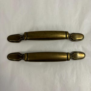 Op de afbeelding: Twee antieke messing kastgrepen. Elke greep heeft een centrale cilindrische handgreep met decoratieve uiteinden. Het messing heeft een warme, verouderde patina. Deze grepen zijn ontworpen voor eenvoudige installatie op laden of kastdeuren.