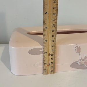 May include: A light pink tissue box with a floral design, approximately 25 cm long. The box features a lily pad and flower motif. A wooden ruler is used to measure the length of the box.