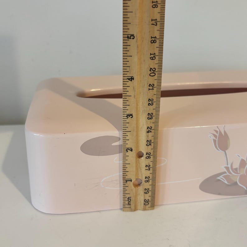 May include: A light pink tissue box with a floral design, approximately 25 cm long. The box features a lily pad and flower motif. A wooden ruler is used to measure the length of the box.