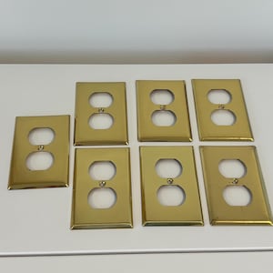 Puede incluir: Siete cubiertas de enchufes eléctricos doradas dispuestas sobre una superficie blanca. Cada cubierta tiene dos aberturas circulares para los enchufes y una forma rectangular. Las cubiertas son de metal brillante.