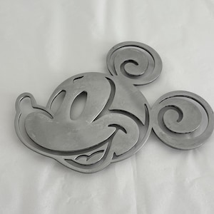 Puede incluir: Un recorte de metal plateado con la cara de Mickey Mouse. El diseño presenta la cabeza, las orejas y la cara sonriente del icónico ratón. El recorte es probablemente un elemento decorativo o parte de un proyecto de manualidades más grande.