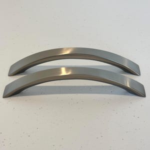 May include: Two brushed nickel cabinet pulls with a curved design. The pulls have a modern, minimalist aesthetic and are likely used for drawers or cabinet doors. The finish is a matte silver color.