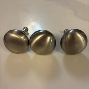 May include: Three brushed nickel cabinet knobs with a spherical design. Each knob has a silver screw attached. The knobs are arranged in a row on a light-coloured surface. These hardware pieces are suitable for drawers and cabinets.