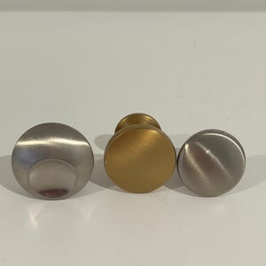 May include: Three round cabinet knobs in brushed metal finishes. One is silver, one is gold, and one is a darker silver. The knobs have a simple, modern design, suitable for drawers or doors.