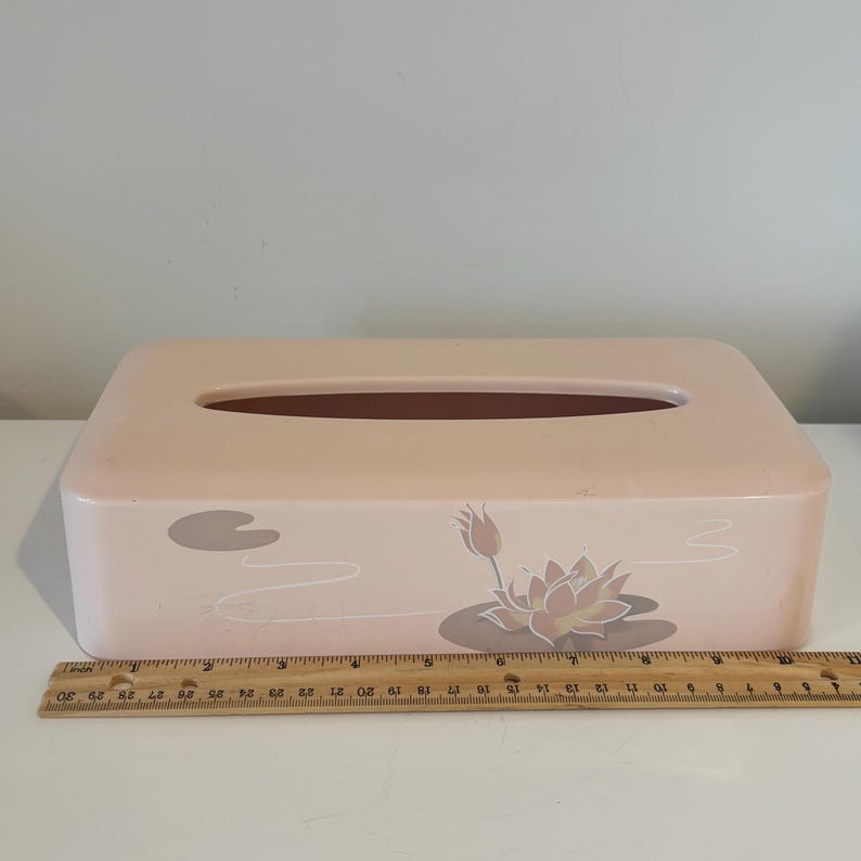 May include: A rectangular tissue box in a soft pink hue, featuring a decorative floral design with lotus flowers and lily pads. The box has a brown opening and measures approximately 25 cm in length.