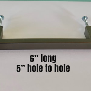 Könnte beinhalten: Ein dunkelgrauer Schrankgriff mit zwei Schrauben. Der Griff ist rechteckig und 15.2 cm lang, mit einem Lochabstand von 12.7 cm. Der Text "6" long 5" hole to hole" ist darunter gedruckt.