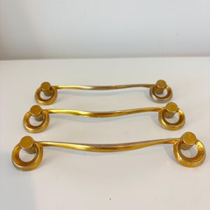 May include: Three gold-colored cabinet pulls with a curved bar and circular ring detail. The pulls are made of metal and have a slightly aged appearance. The hardware is designed for furniture and home decor.