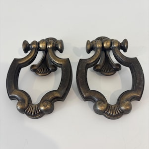 May include: Two antique bronze-toned drawer pulls. Each pull features a decorative ring design with a central handle and ornate detailing. The pulls are made of metal and are likely for furniture restoration or decorative use.