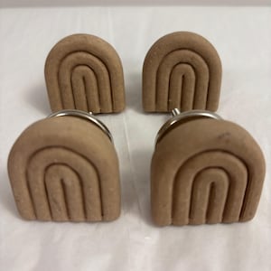 Peut inclure: Quatre boutons de meuble bruns en forme d'arc-en-ciel avec des fixations argentées. Les boutons sont faits d'un matériau texturé et sont disposés en carré. Le motif arc-en-ciel présente plusieurs lignes courbes, créant un effet superposé. Pour tiroirs ou armoires.