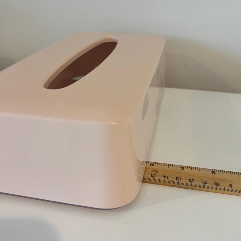 May include: A rectangular, light pink tissue box with an oval opening on top. The box is approximately 38 cm long. The tissue box is on a white surface.