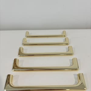 May include: A set of five gold-colored cabinet pulls arranged in a row. Each pull has a rectangular bar with curved ends. The pulls are made of a shiny, reflective material.