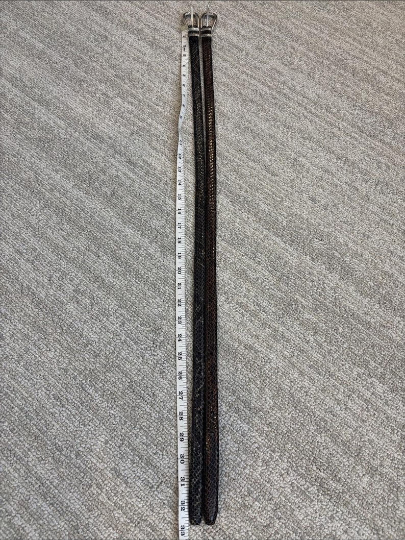 May include: Two dark brown leather belts with silver buckles, each approximately 33 inches long, are laid side by side. The belts have a woven texture and are positioned vertically against a gray textured background.